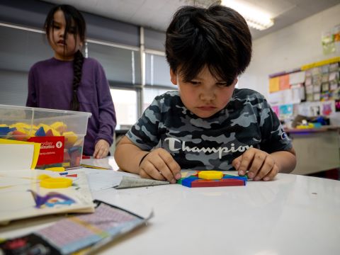 A young student focuses on completing a colorful puzzle at his desk in a bright classroom.
