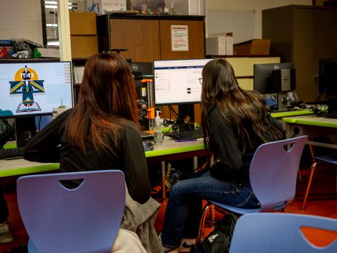 Two students are sitting at desks, focused on their computers while working on a project together.