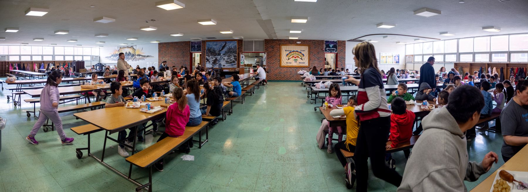 A busy school cafeteria filled with students eating lunch and chatting at various tables.