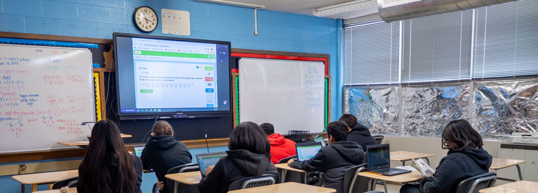 Students at desks in a classroom, with graphing problems on a large screen and a school clock hanging above.