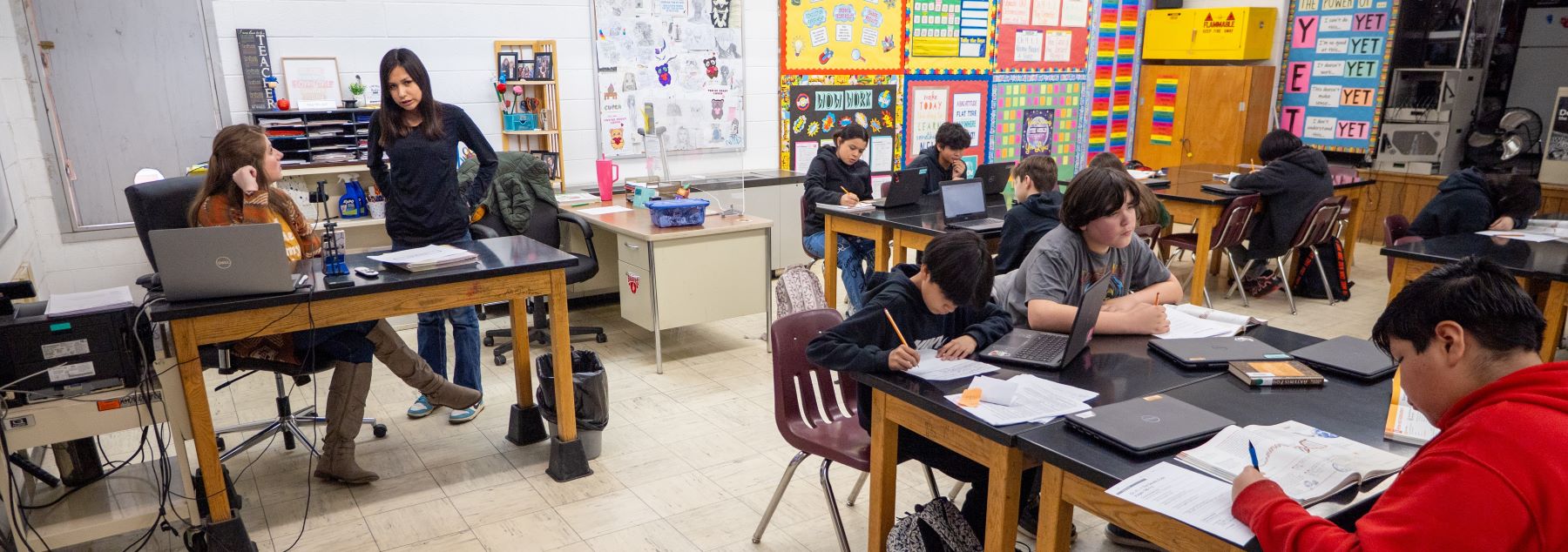 Students sitting at desks with laptops in a classroom, working on their assignment while a teacher works with a student at her desk.