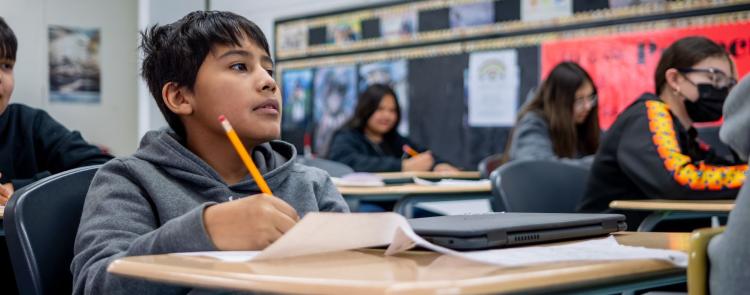 A student sits at a desk, holding a pencil, focused on his class work.